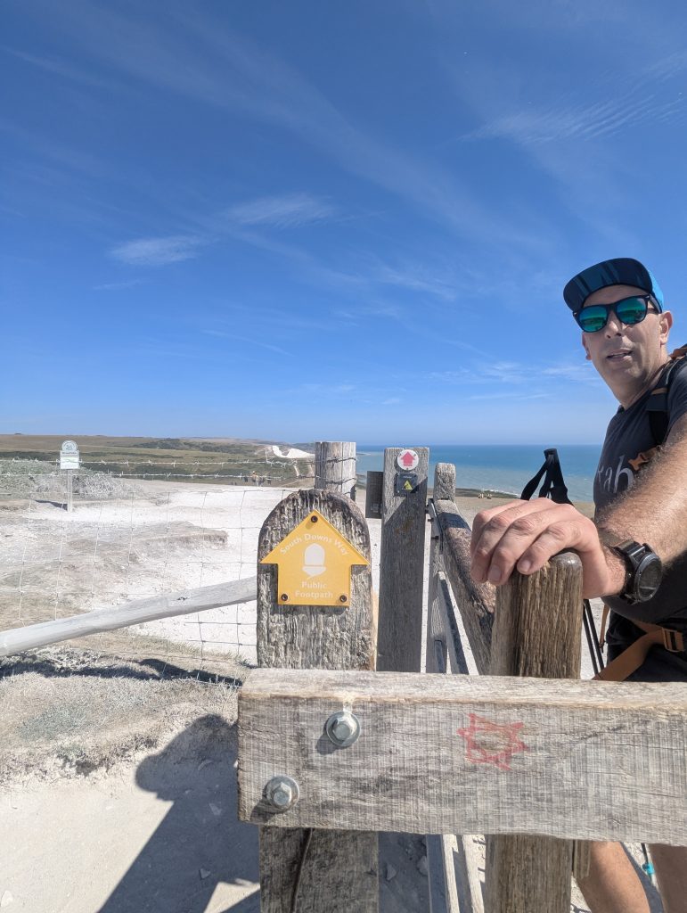Hiker Terry leaning on a wooden fence stile with a yellow South Downs Way public footpath acorn arrow signpost overlooking the coastal hills and sea.