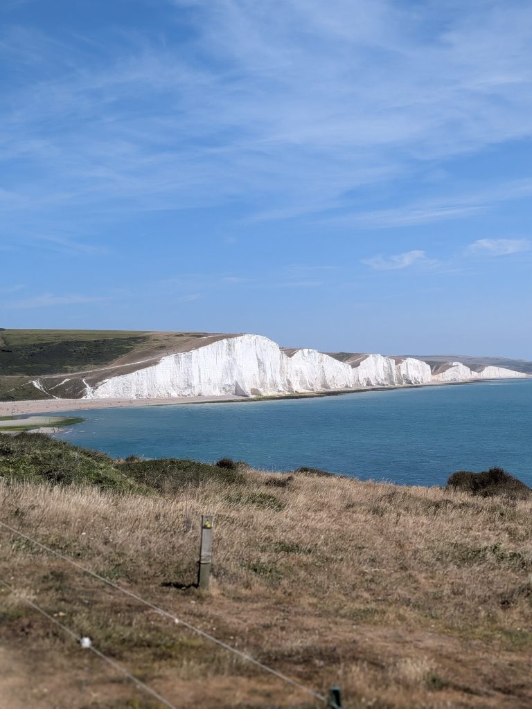 Wide landscape view across a calm blue sea toward the glittering white chalk cliff faces of the Seven Sisters rising above the dry grassland of Cuckmere Haven.