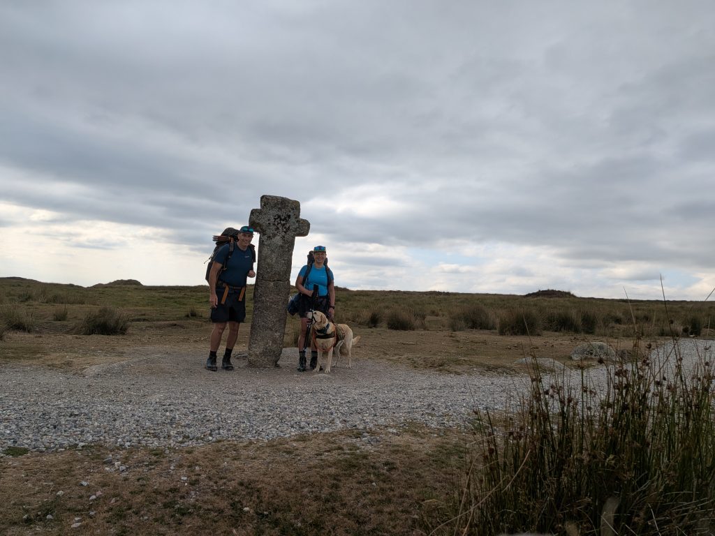 Terry and Syla wearing backpacks standing beside ancient granite Siward's Cross on a gravel path with Poppy the yellow Labrador cross on a lead.