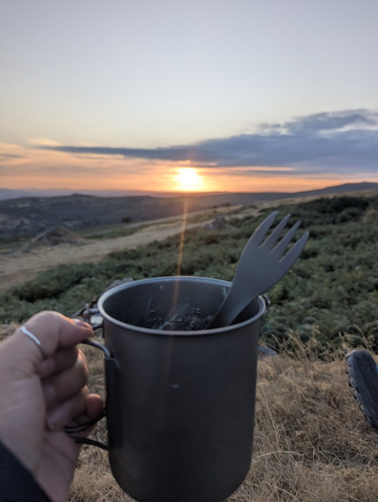 Hand holding a titanium camping mug with a spork against a golden sunset over Swelltor Quarry on Dartmoor.
