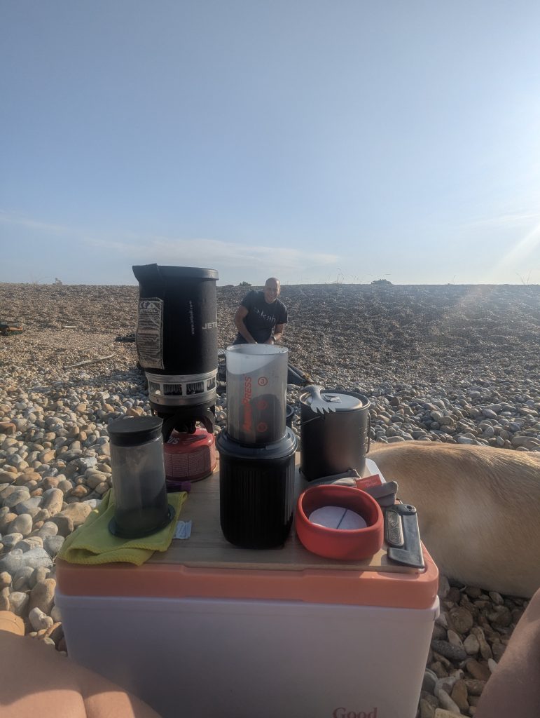 Close-up of an Aeropress coffee maker and Jetboil stove on a camp cooler, with a male hiker and yellow Labrador cross Poppy in the background on Dungeness shingle.