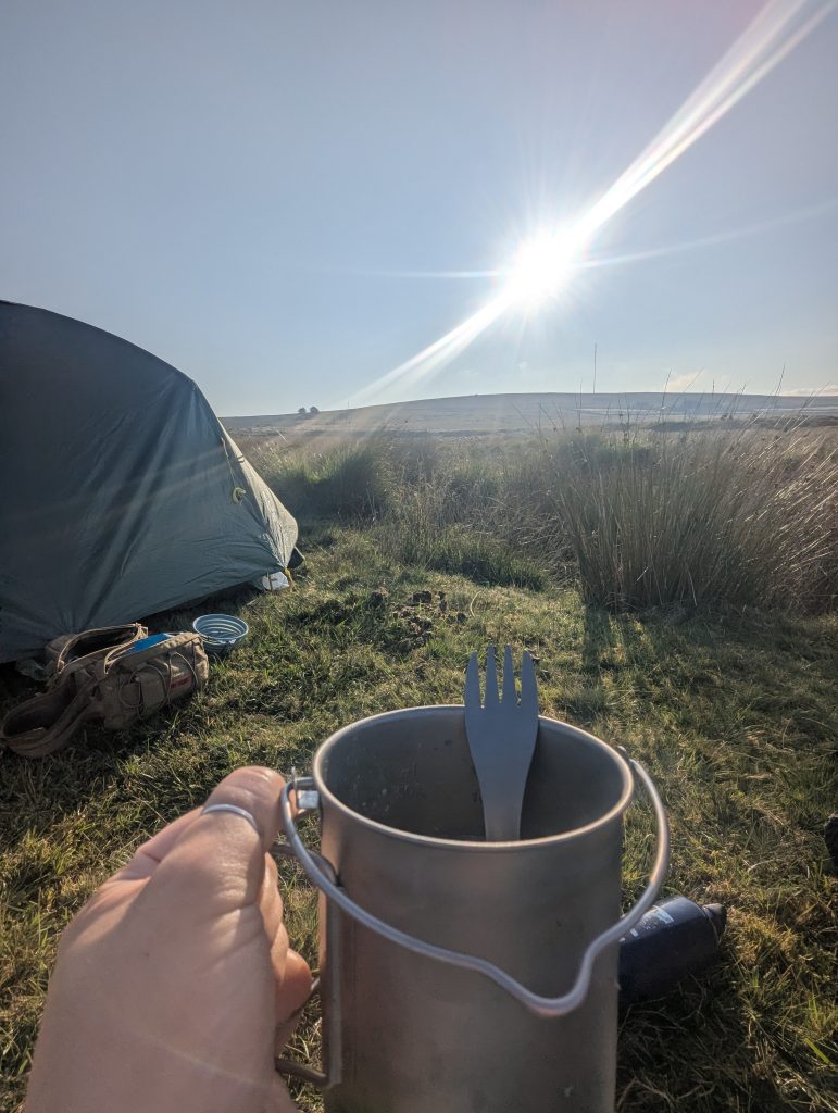 Hand holding a titanium camping mug with a spork inside, in front of a green tent set up on grassy moorland under bright morning sunlight.