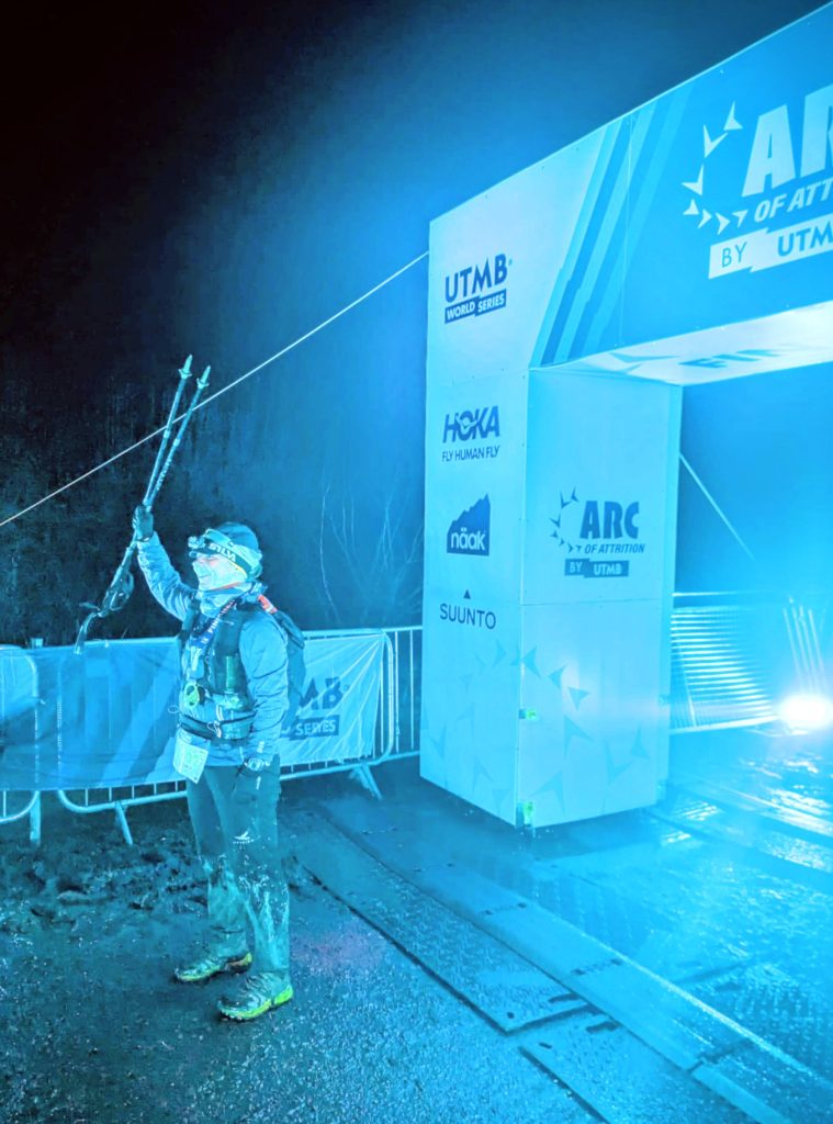 A muddy ultra-runner celebrating at the night finish line of the Arc of Attrition race, holding up trekking poles. The runner is wearing Harrier Dartmoor waterproofs covered in grime.