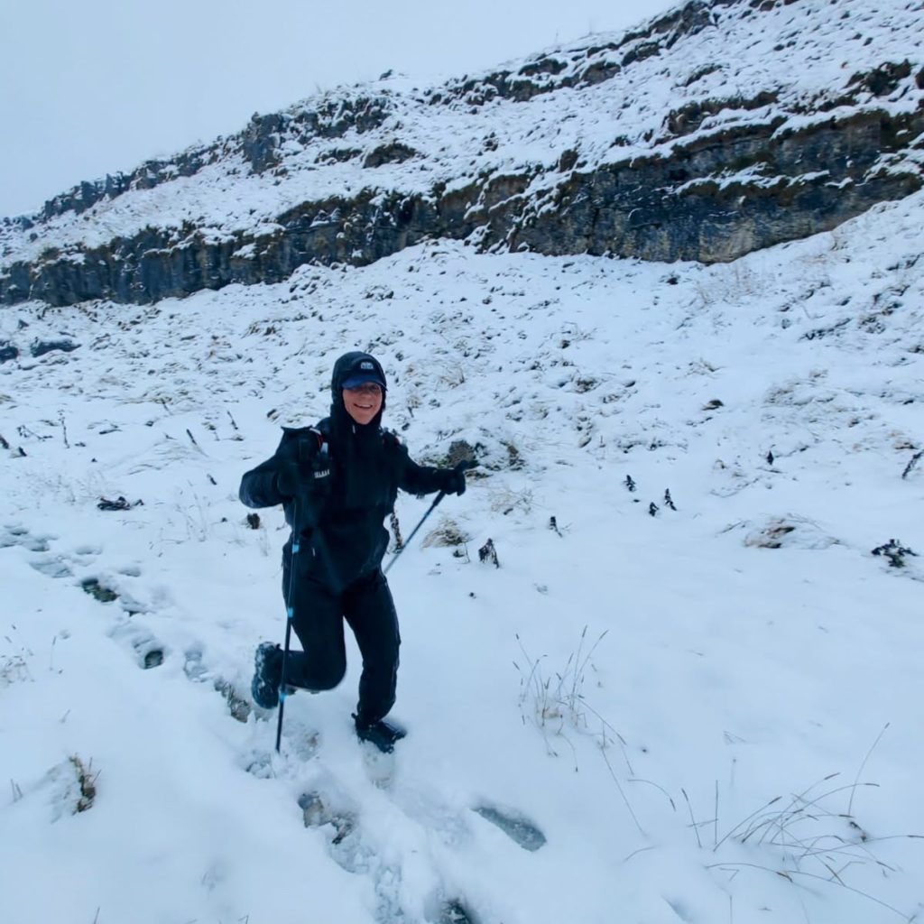 A female athlete running uphill through deep snow on a mountain path, wearing black Harrier Dartmoor waterproof overtrousers and a matching jacket. She is using trekking poles.