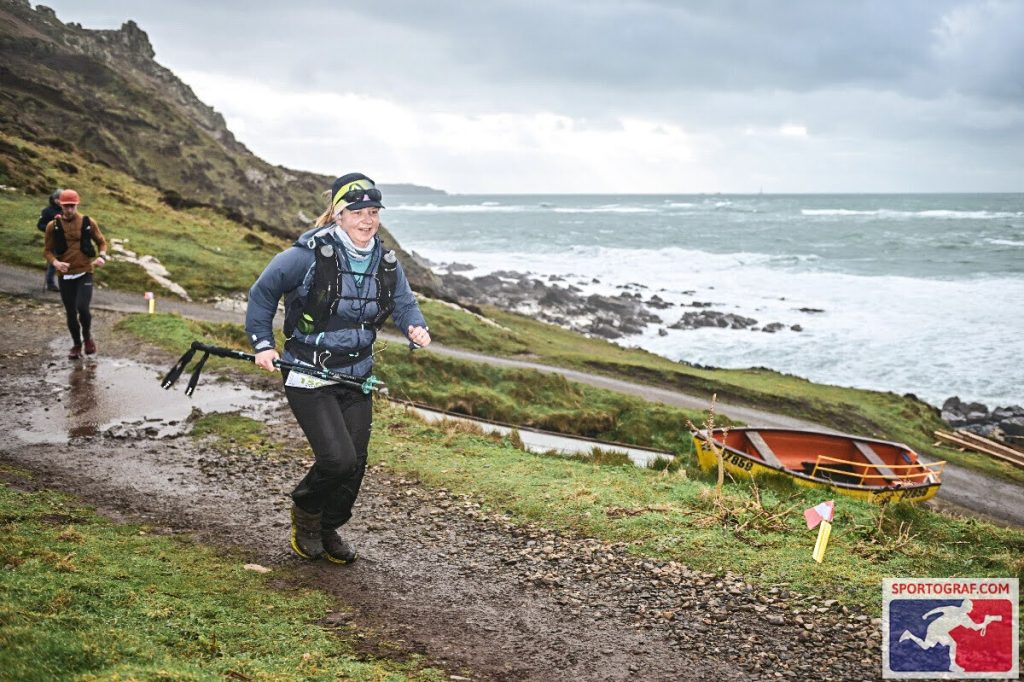 Female ultra-runner wearing black Harrier Dartmoor waterproof trousers and race gear, navigating a muddy coastal trail beside rough seas during the Arc of Attrition.