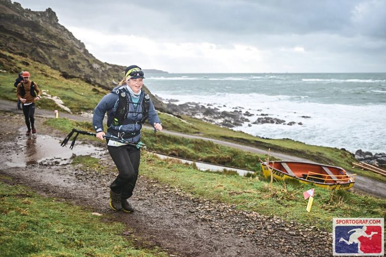 Female ultra-runner wearing black Harrier Dartmoor waterproof trousers and race gear, navigating a muddy coastal trail beside rough seas during the Arc of Attrition.
