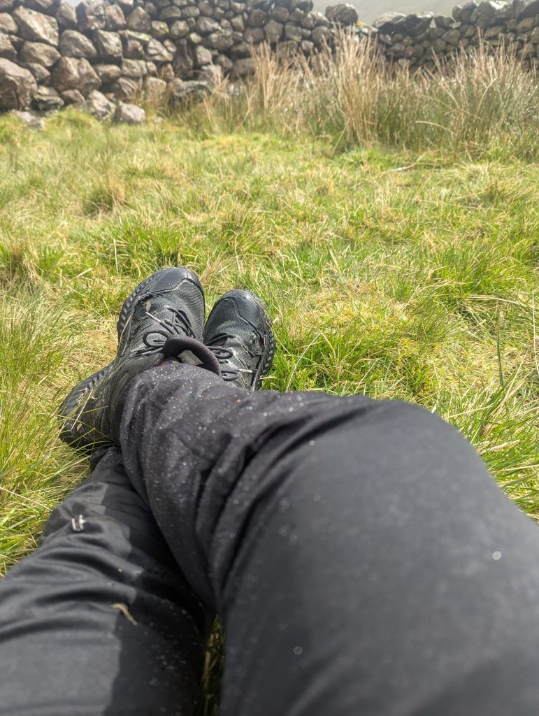 A close-up first-person view looking down at the fabric of the black Harrier Dartmoor trousers. Water is beading on the surface after rain, illustrating the DWR performance.