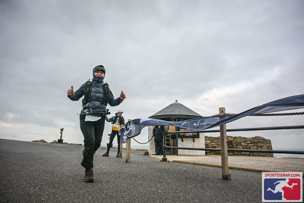 Female ultra-runner giving a thumbs-up while wearing black Harrier Dartmoor waterproof trousers and jacket at a coastal race checkpoint during the Arc of Attrition.