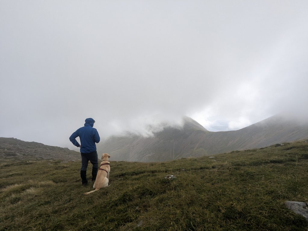 A hiker wearing a blue jacket and a yellow Labrador sit on a grassy mountain crest looking out toward atmospheric clouds moving over adjacent peaks.
