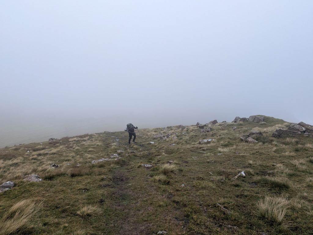 A lone hiker with trekking poles and a heavy rucksack walks along a grassy mountain ridge under a dense blanket of grey cloud and low visibility.