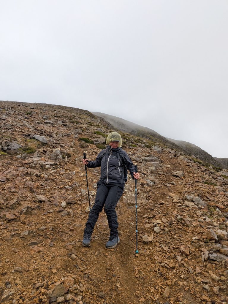 Hiker in waterproof jacket and head cover descending gravel mountain trail in fog wearing Inov-8 Roclite Pro Mid GTX boots.
