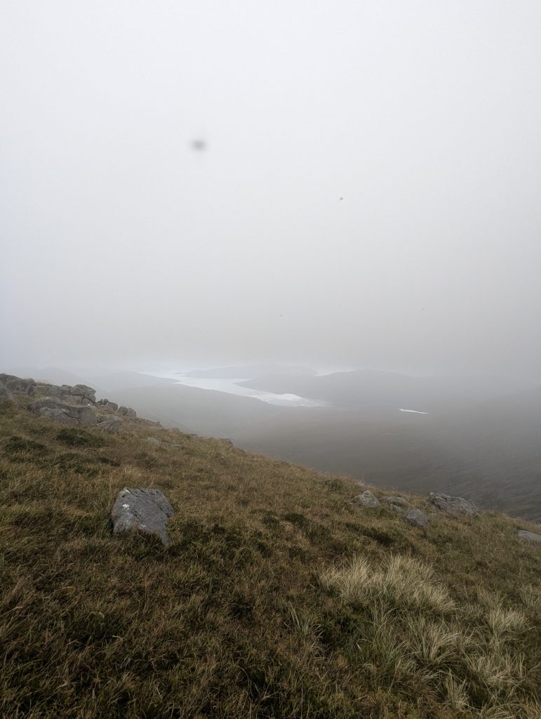 View from a grassy mountain ridge looking down into a valley where thick mist partially obscures a loch in the distance.