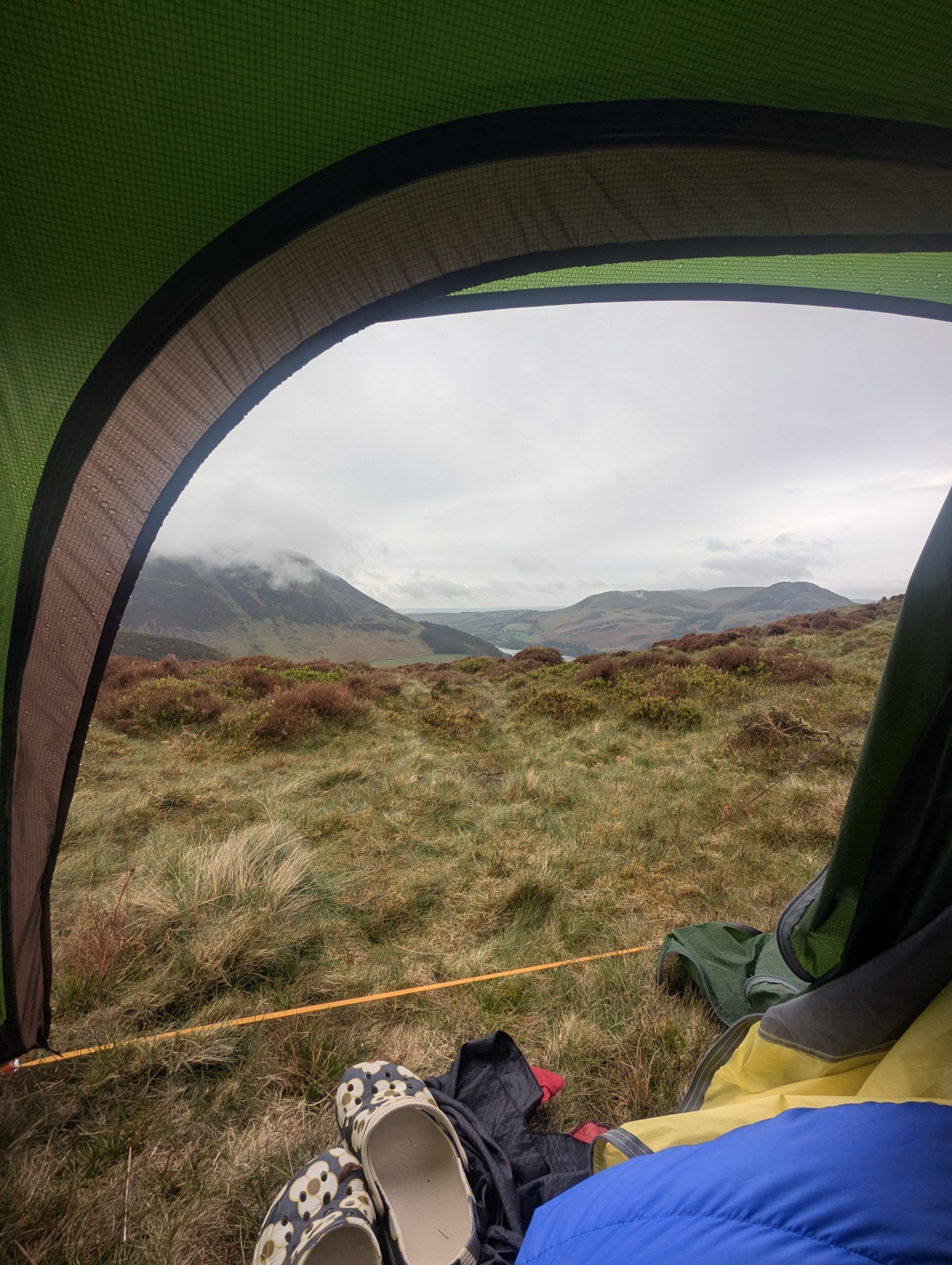 View from inside a green tent looking out over a cloudy mountain valley, with a pair of black and cream patterned Orla Kiely x Regatta slip-on clogs resting on the grass next to a blue sleeping bag.