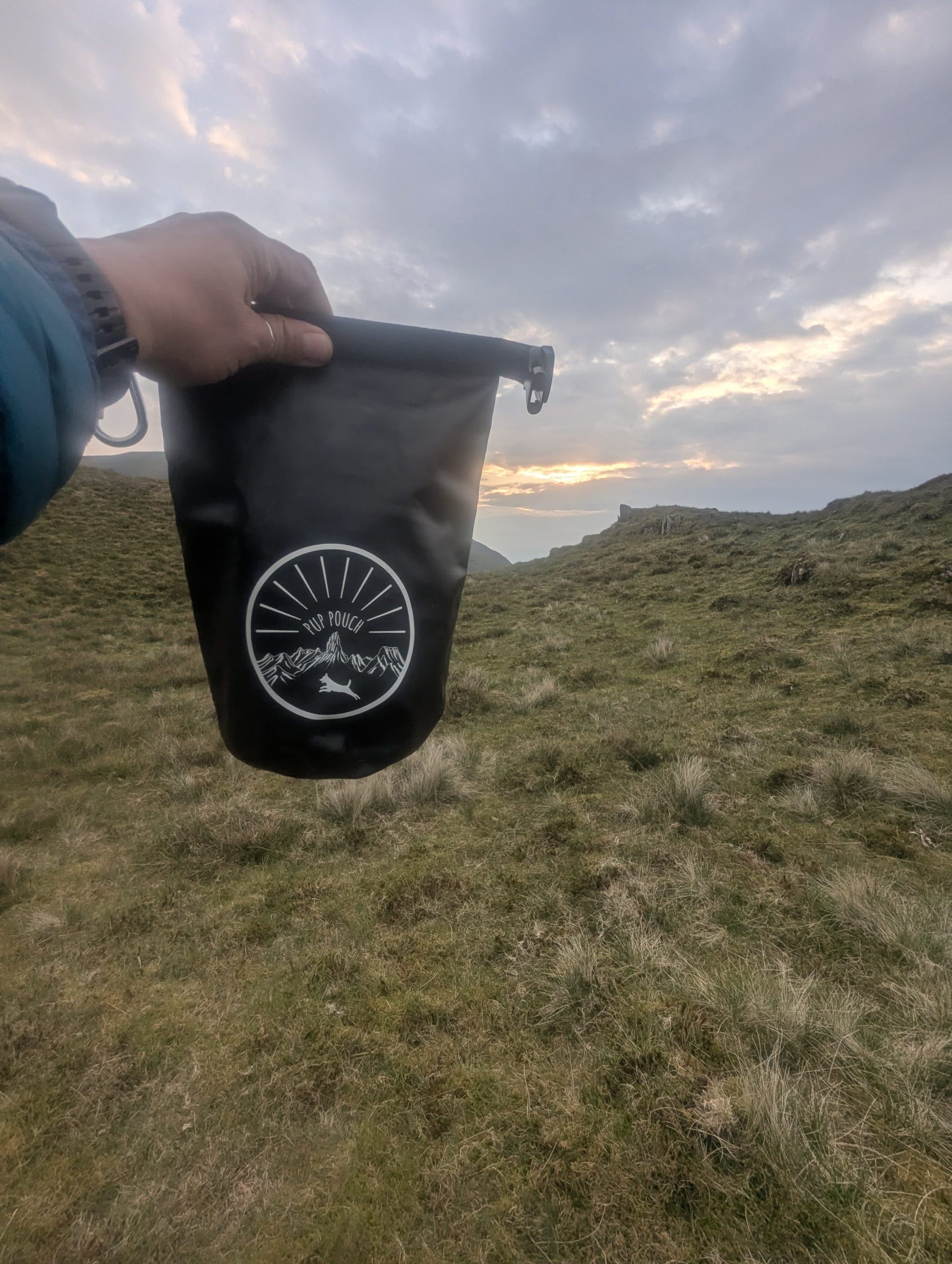 Essential Multiday Hike Gear List Item - Hand holding a black waterproof Pup Pouch dry bag up against a dramatic sunset sky over a grassy mountain ridgeline in Eryri / Snowdonia.