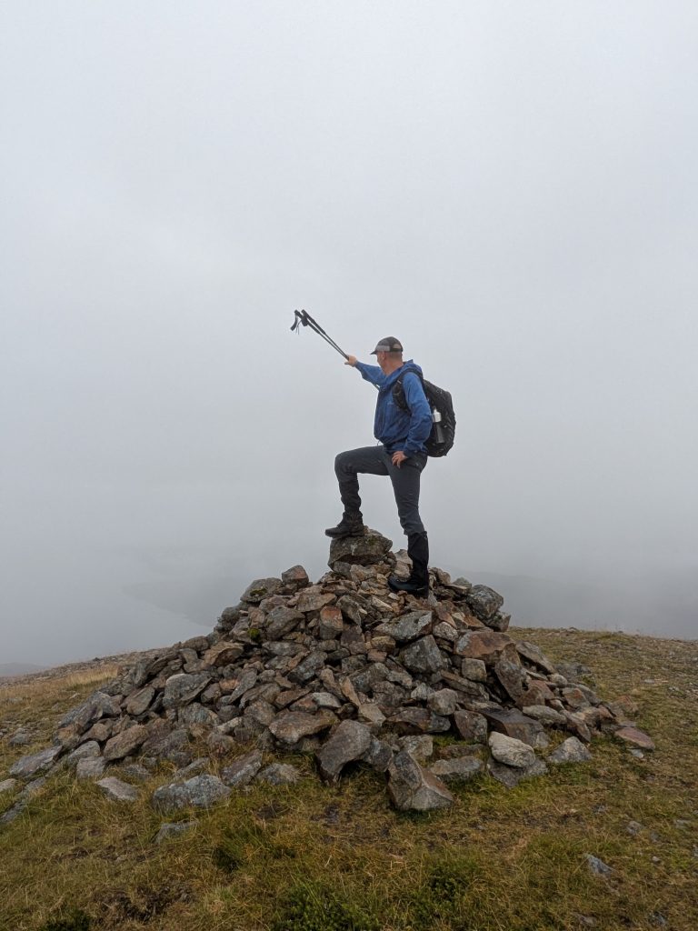 Hiker with a dark backpack and trekking poles standing atop a large, loose stone cairn, pointing forward with their poles into dense, low-visibility fog.