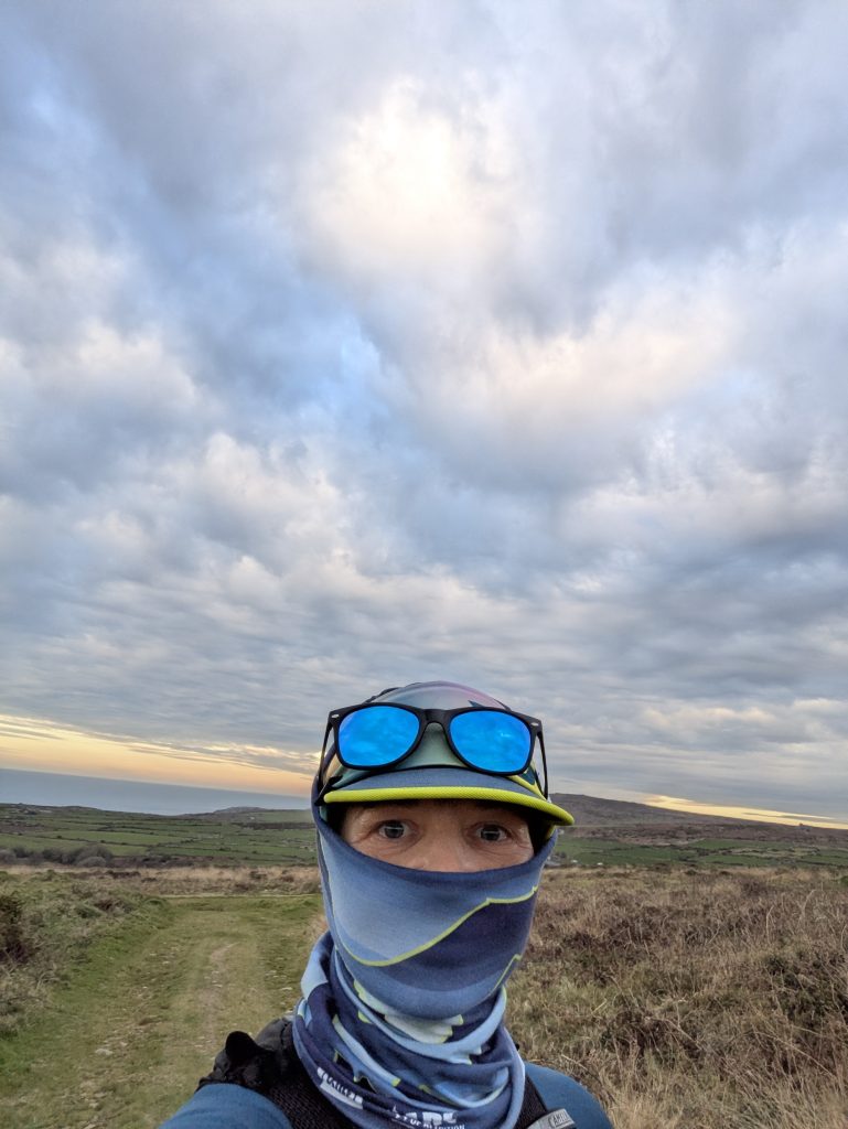 Hiker wearing a blue and yellow VAGA 13 Valleys Ultra Edition lightweight running cap with a blue mountain-patterned snood pulled up over the brim and sunglasses to block heavy wind on an multiday hike.