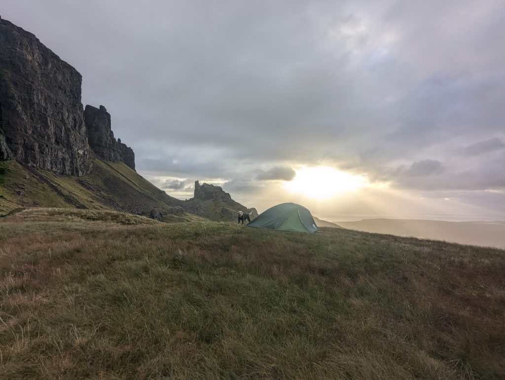 First-person view from inside a green wild camping tent looking past a pair of patterned camp clogs towards a dramatic golden hour sunset over a sweeping, grassy mountain valley.