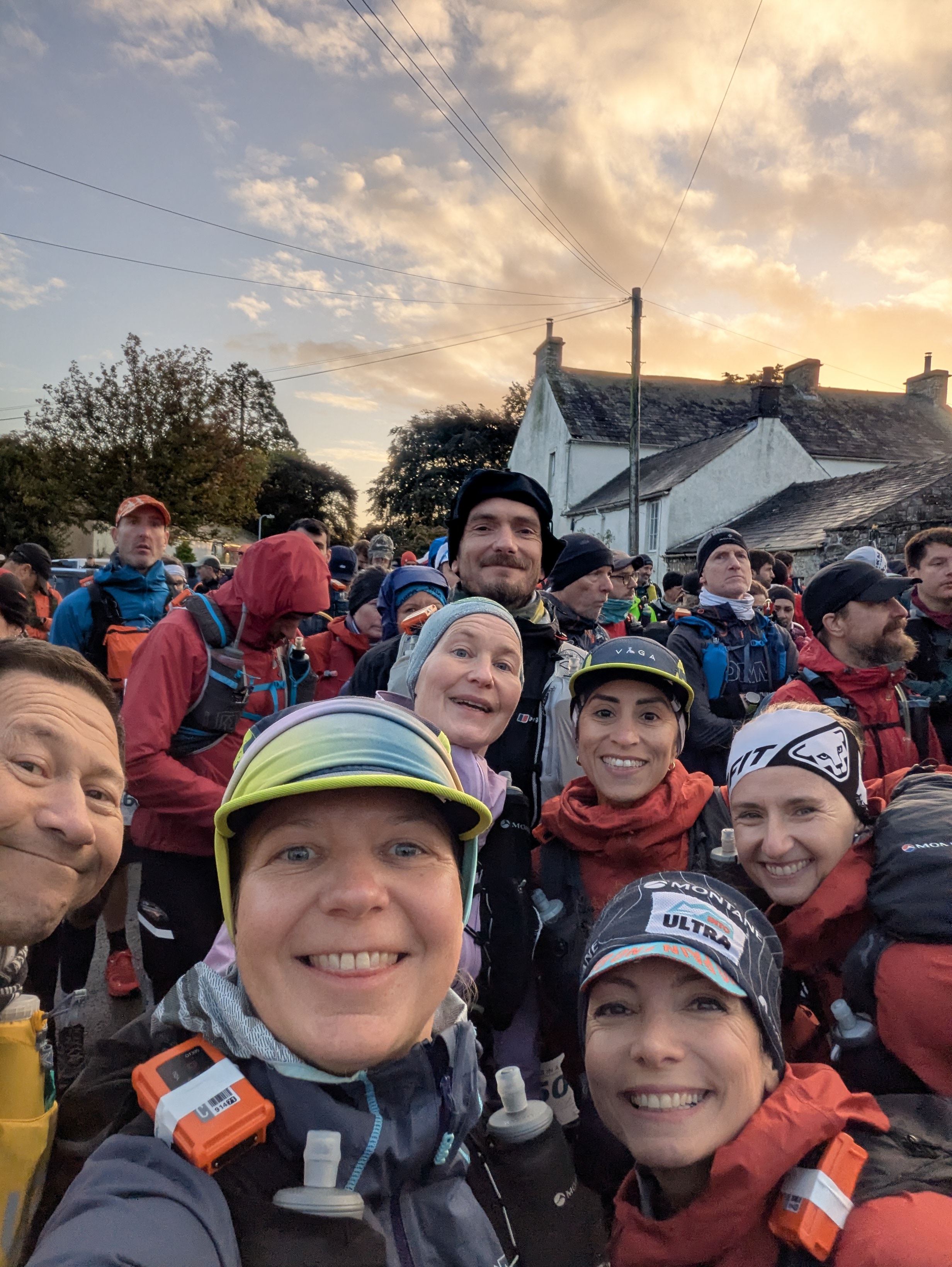 Group of smiling trail runners taking a sunrise selfie at the start line of a Lake District ultra marathon.