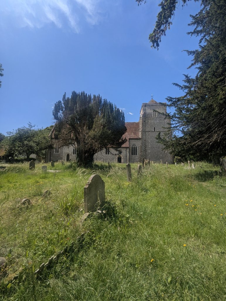 A historic stone church with a square tower and traditional graveyard surrounded by green grass and mature trees along the trail in Bridge, Kent.