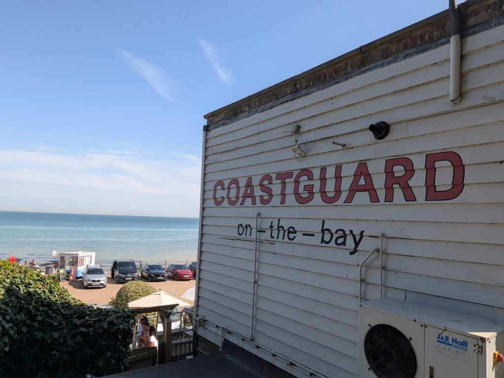 White wooden exterior of The Coastguard pub with large red lettering reading COASTGUARD on-the-bay, overlooking the beachfront car park and sea.
