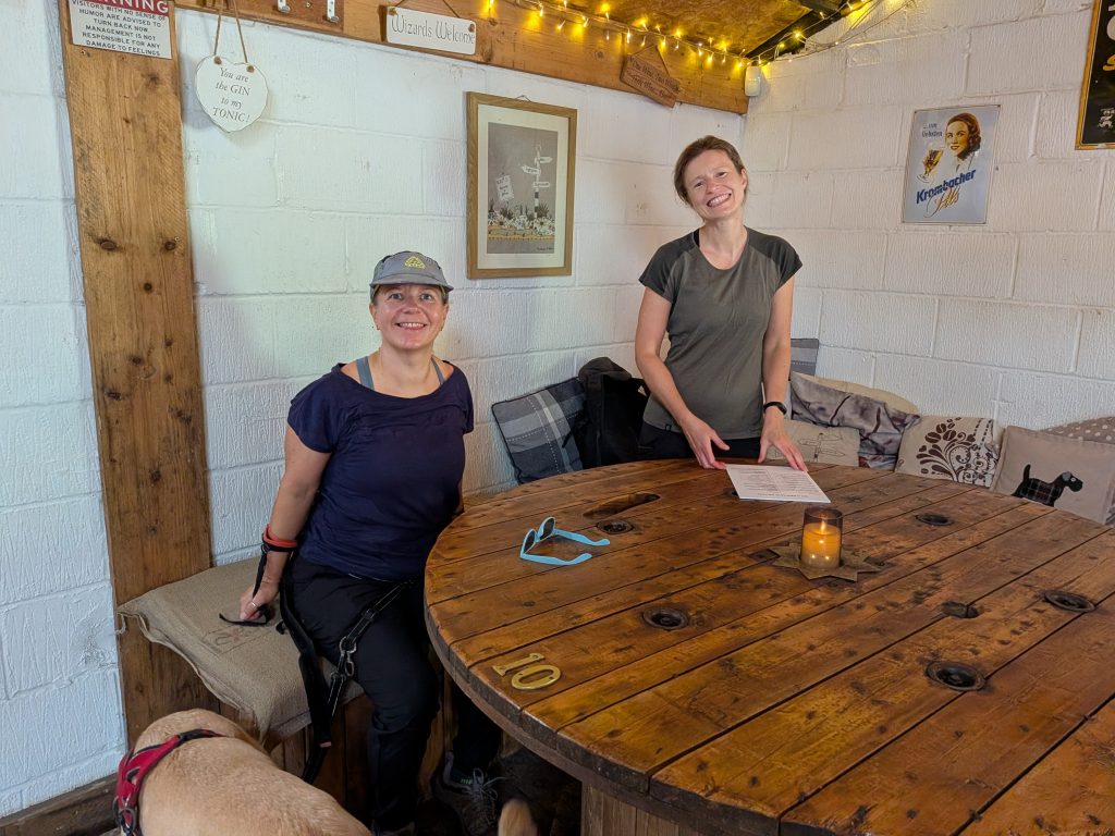 Syla and a hiking companion (Aga) smiling inside a rustic pub, sitting at a large wooden cable spool repurposed as a table, with yellow Labrador Poppy resting beside them.