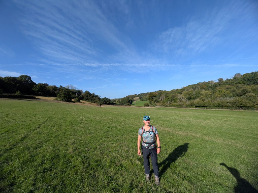 A female hiker with backpack standing in a lush green valley framed by wooded hills under a bright blue sky