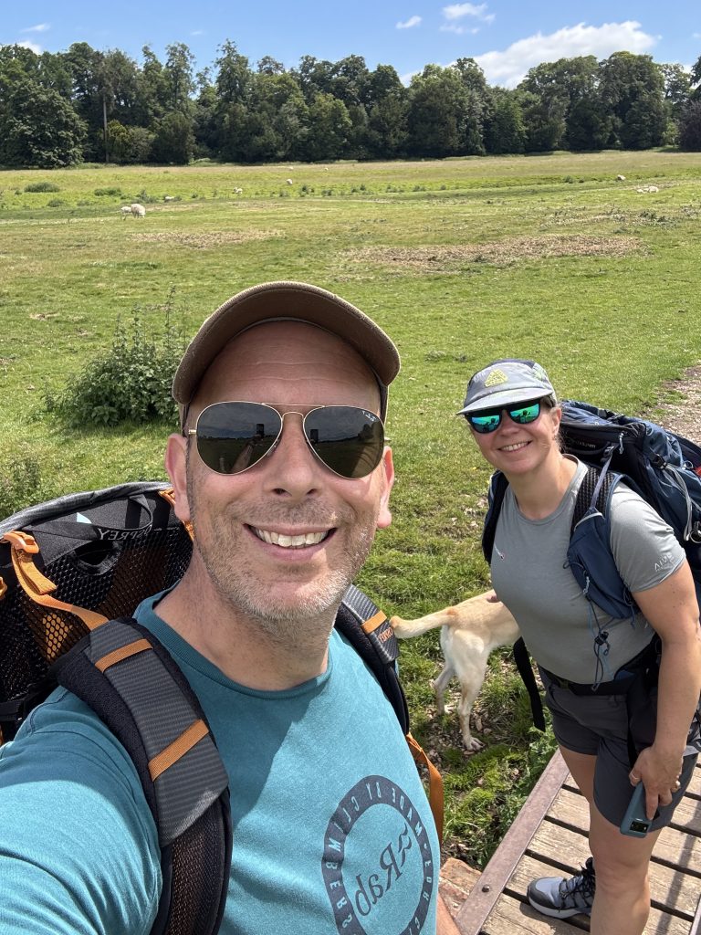 A smiling couple taking a selfie on a wooden footbridge over an Elham Valley pasture with a dog and sheep grazing in the background.