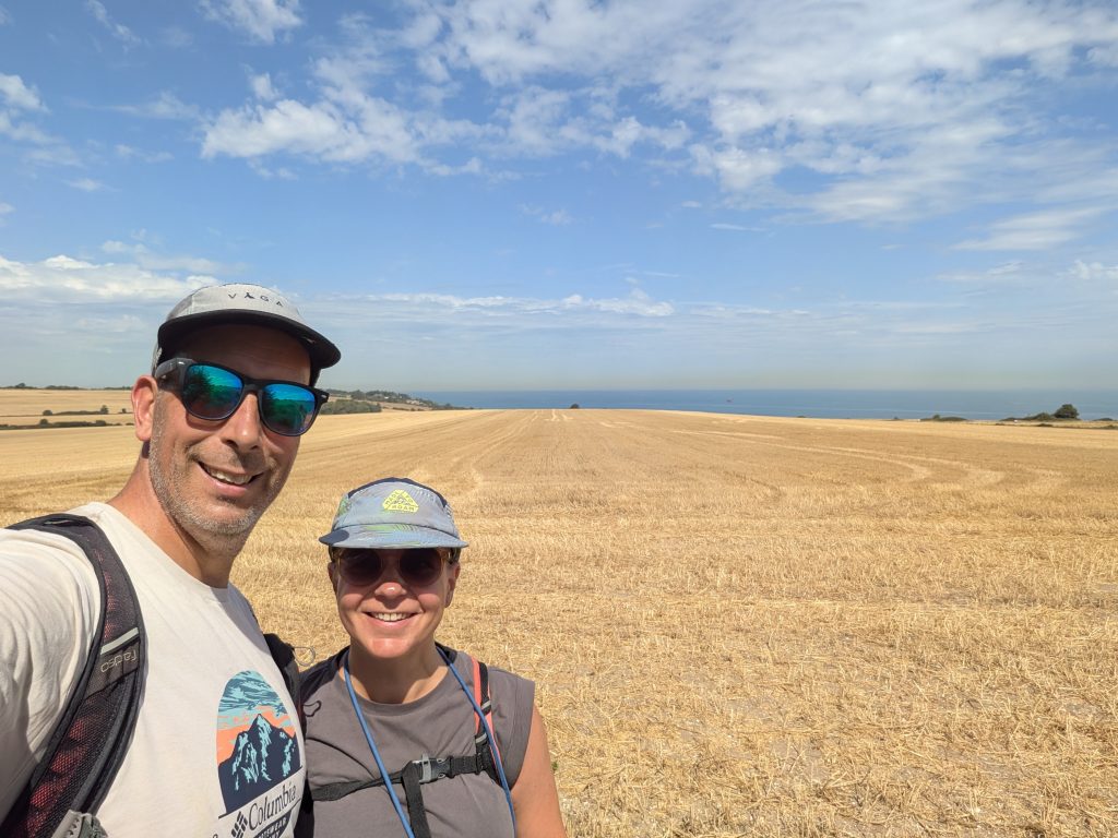 A selfie of a man and woman smiling in front of a vast golden field with the ocean in the background.