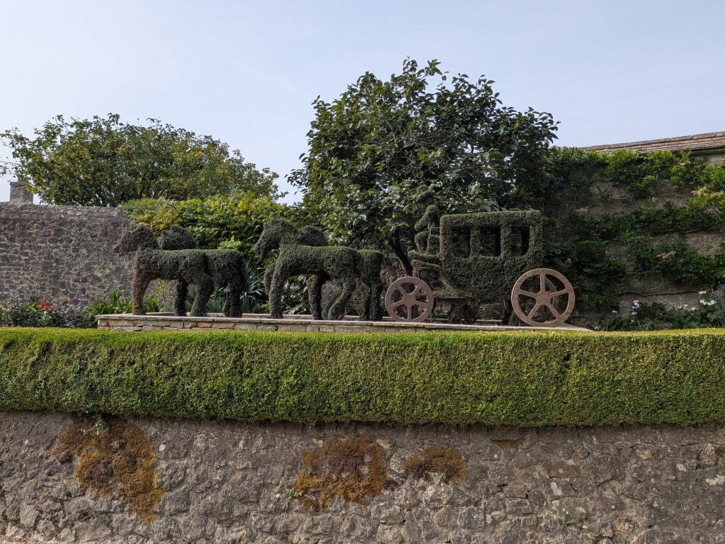 A meticulously shaped topiary hedge depicting two horses pulling a traditional carriage with a driver, set against a stone wall and mature trees.