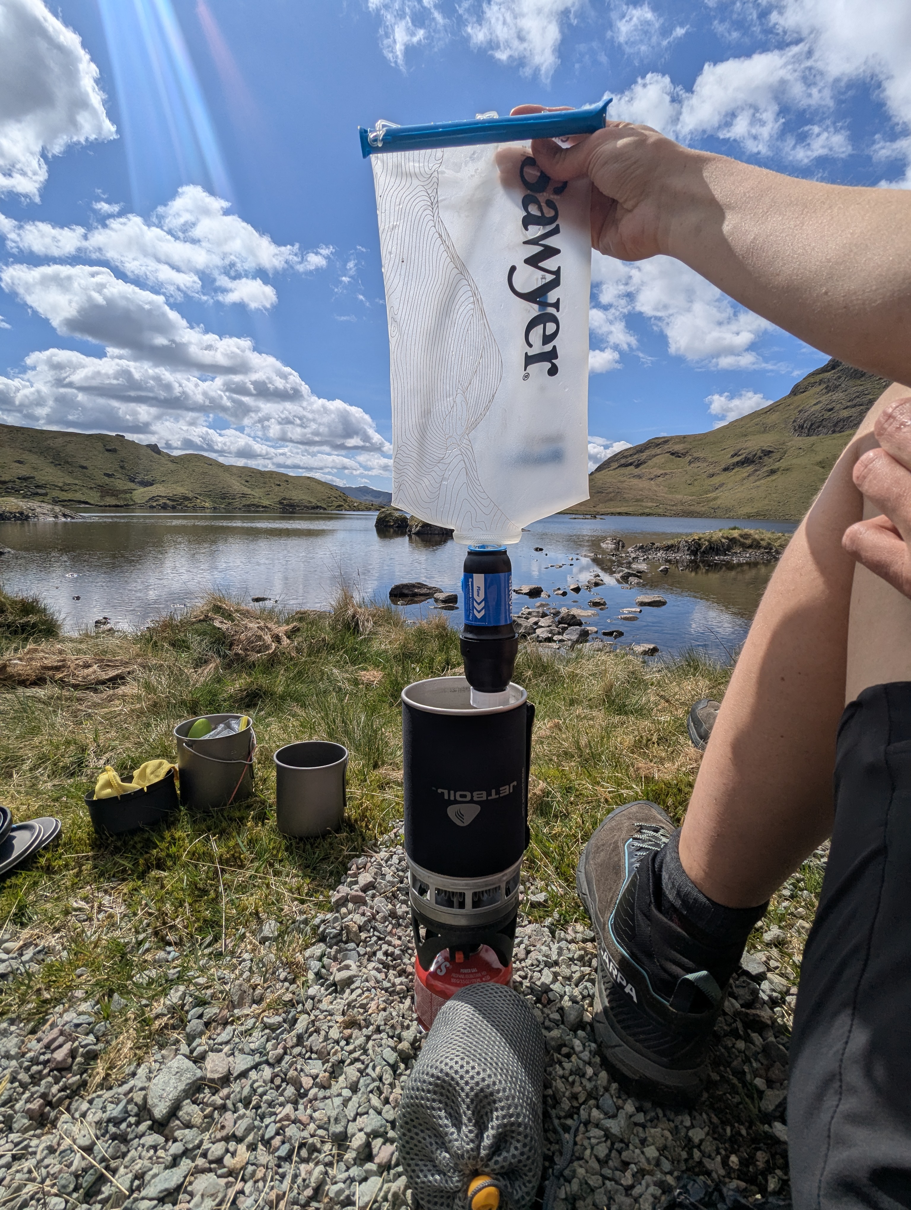 Hiker wearing Scarpa trail shoes filtering lake water through a Sawyer Squeeze and Cnoc bladder directly into a Jetboil PCS cup on a grassy lakeshore.