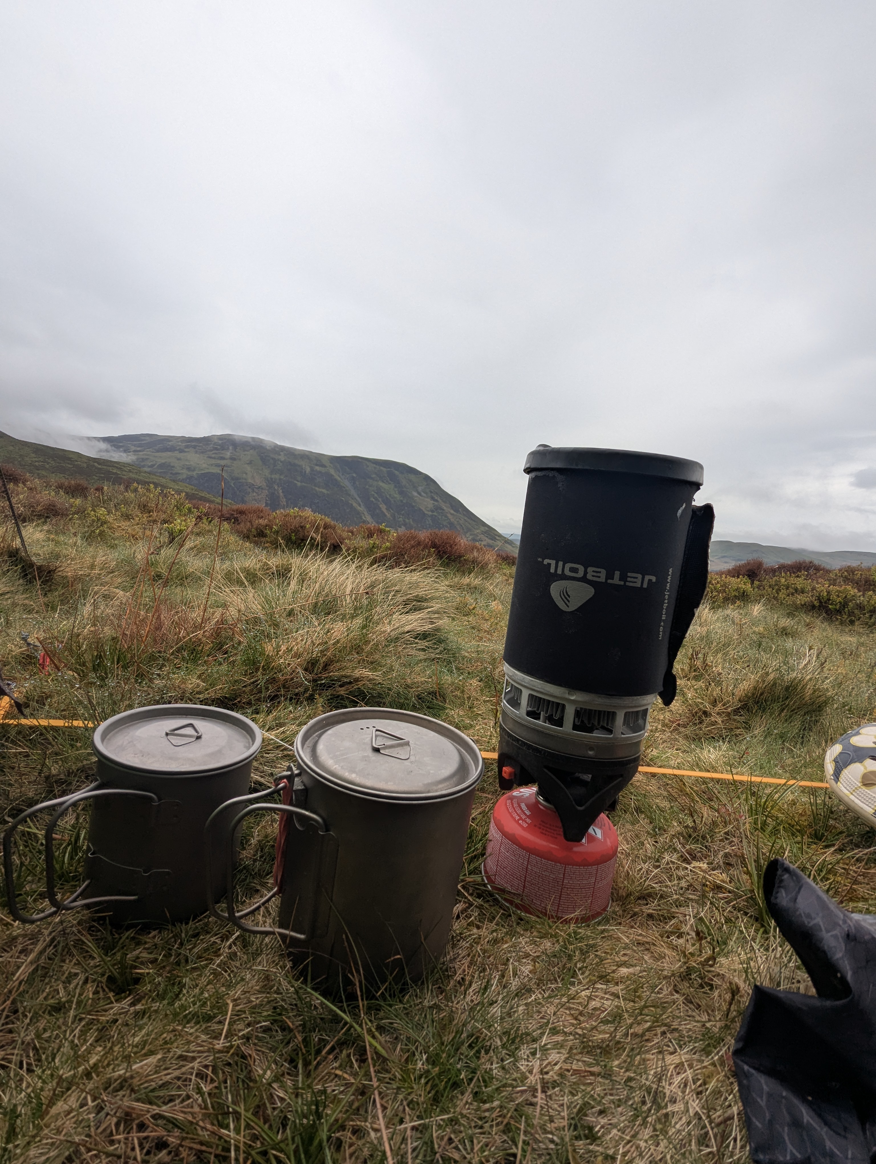 Jetboil PCS stove leaning slightly on uneven mountain grass next to two grey titanium camping mugs with a steep mountain ridge in the background.