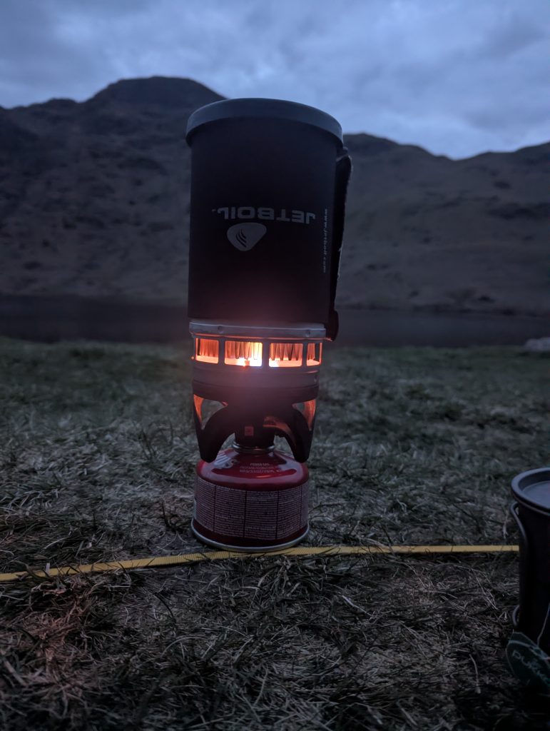 Jetboil PCS operating at dusk with the corrugated metal FluxRing glowing bright orange from the burner heat, sitting on dark grass beside a mountain lake silhouette.