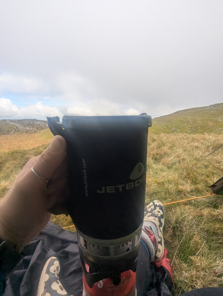Hiker's hand holding a steaming black 2006 Jetboil PCS cup with a worn neoprene sleeve over a burner on a grassy mountain plateau in Eryri.