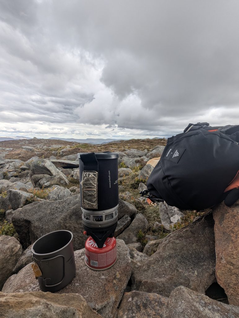 Jetboil PCS operating at dusk with the corrugated metal FluxRing glowing bright orange from the burner heat, sitting on dark grass beside a mountain lake silhouette.