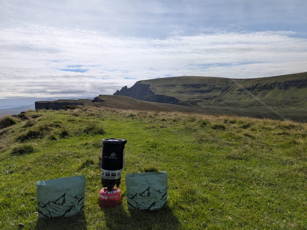 Jetboil PCS on a red gas canister flanked by two green Food Force dehydrated beef pasta meal pouches on a bright green grassy cliff edge with steep mountain drop-offs behind.