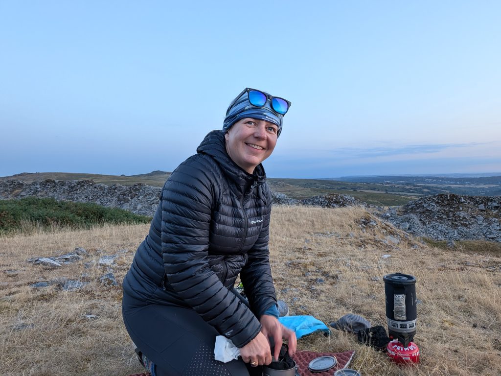 Female hiker wearing a black Montane down jacket and blue sunglasses smiling while setting up a Jetboil PCS on a red Primus gas canister during a dry moorland wild camp.