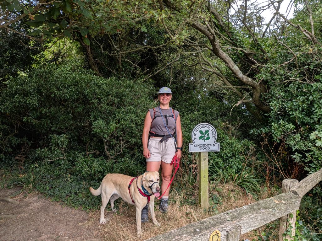 A female hiker smiling with her yellow Labrador next to a National Trust sign for Kingsdown Wood.