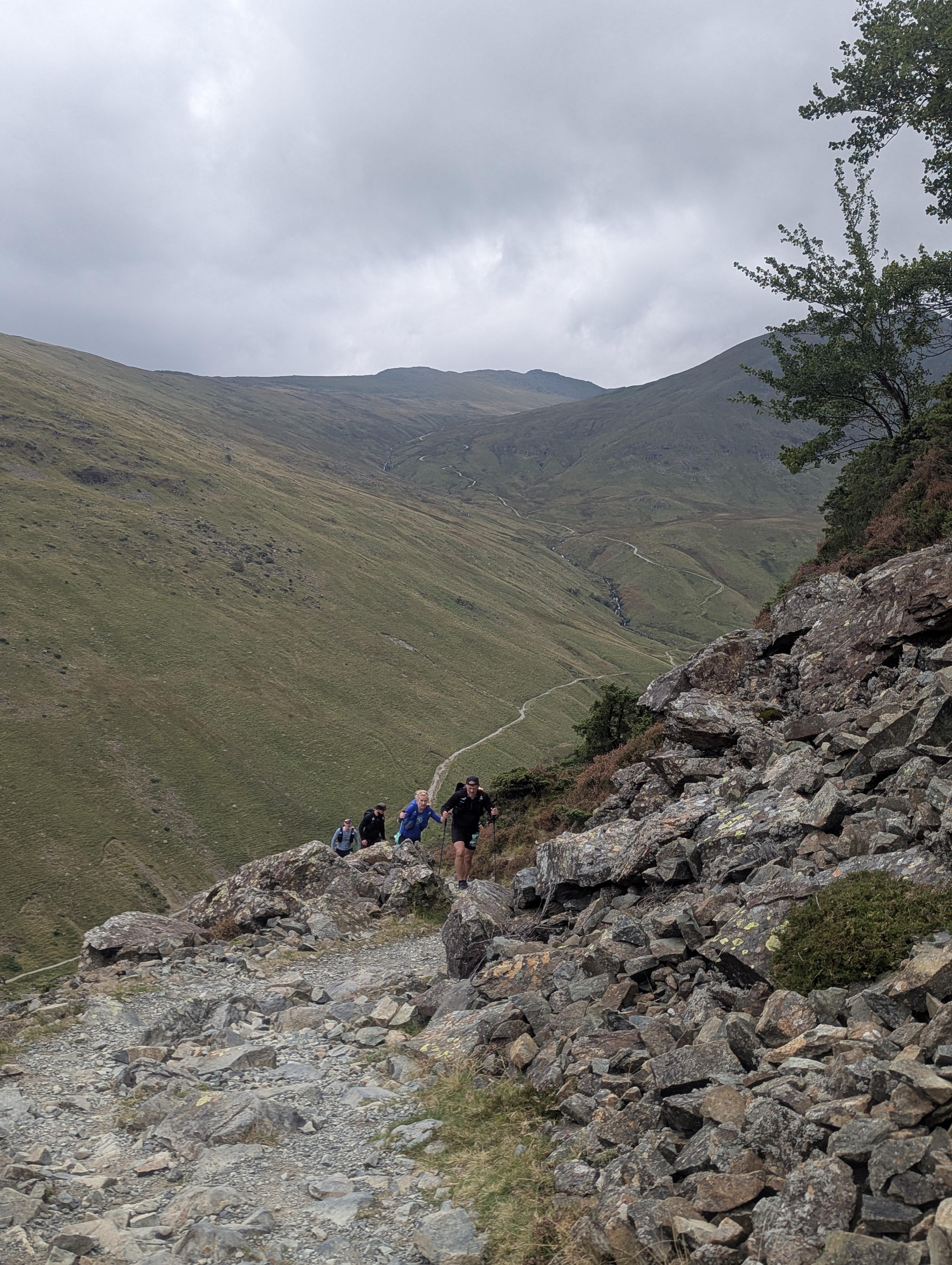 Group of ultra runners climbing a steep, rocky mountain path in the Lake District under overcast skies.