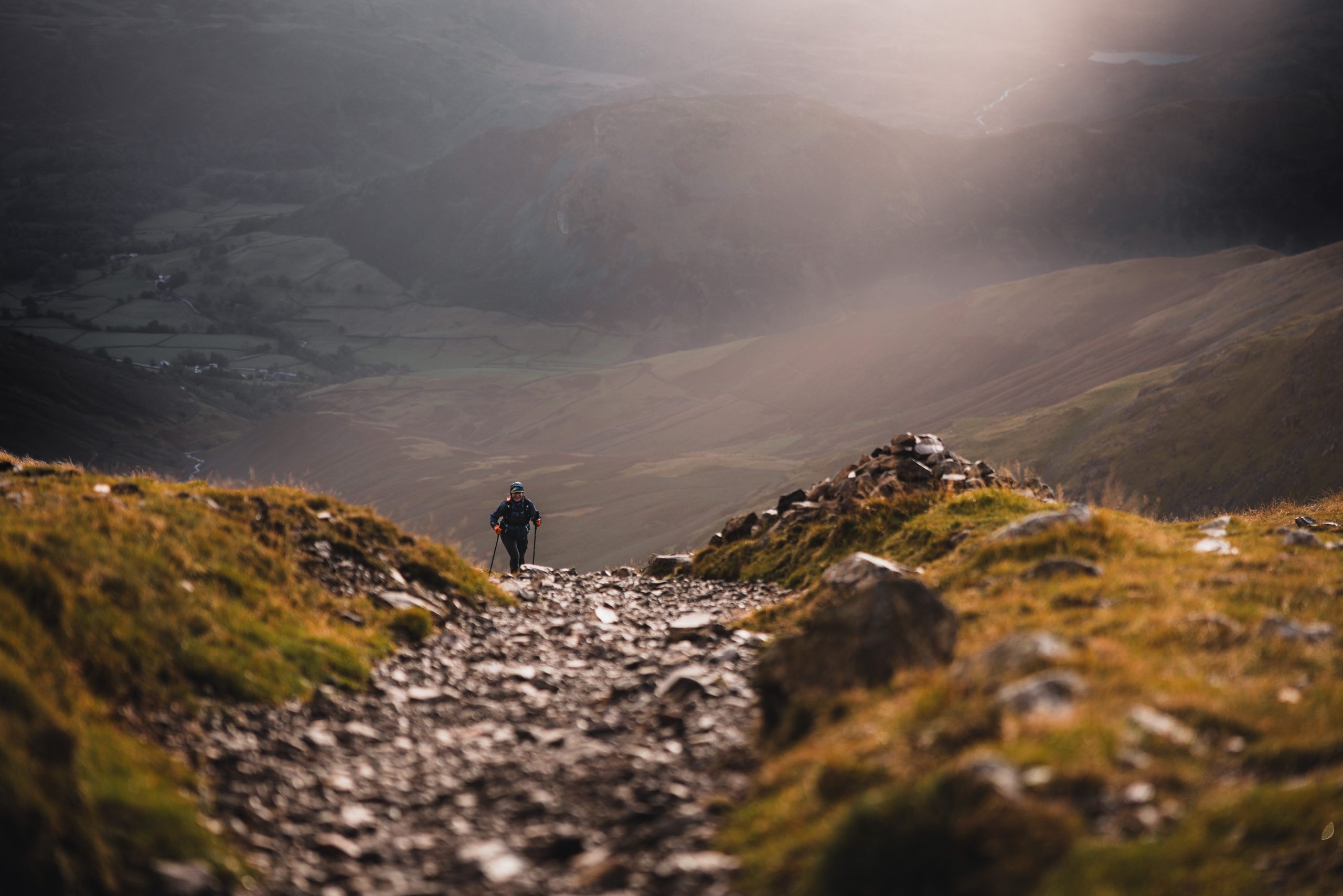Solo ultra runner navigating a steep, rocky descent in the Lake District fells during golden hour sunlight.
