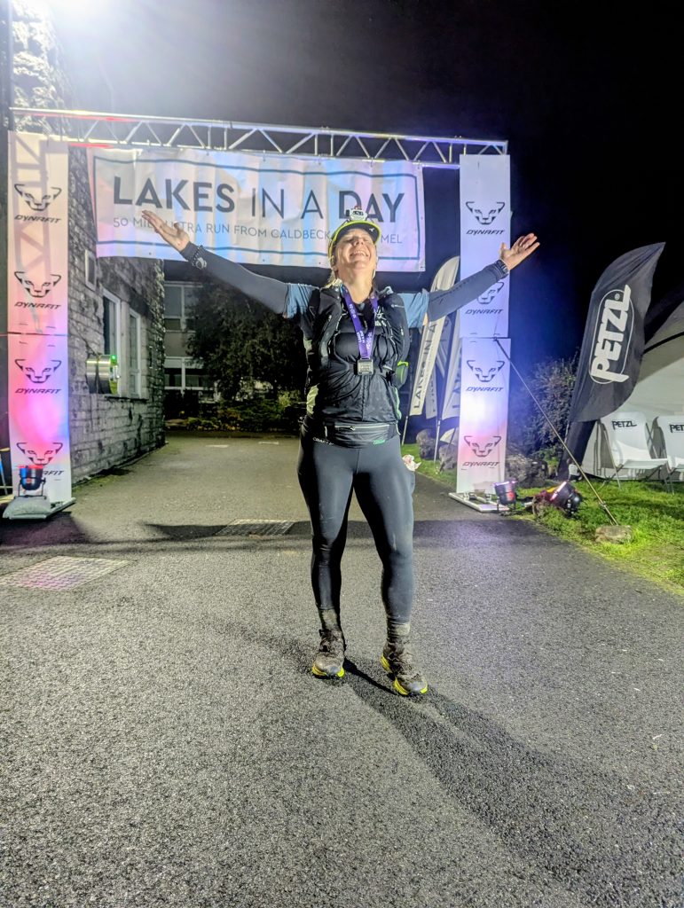 Exhausted yet joyful ultra runner raising arms in celebration under the finish arch at Cartmel.