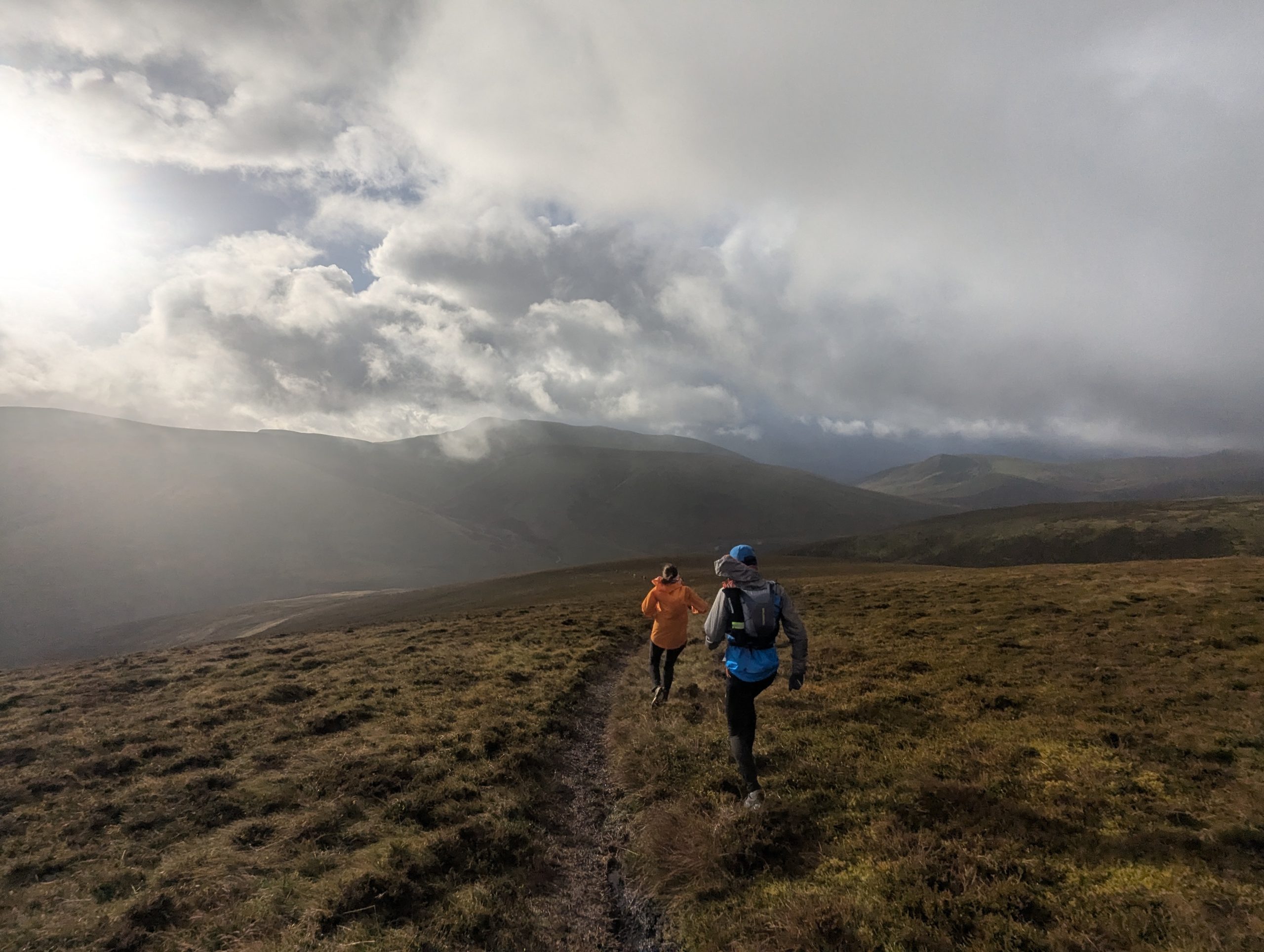Two trail runners moving across open, grassy fell terrain under an expansive cloudy sky.