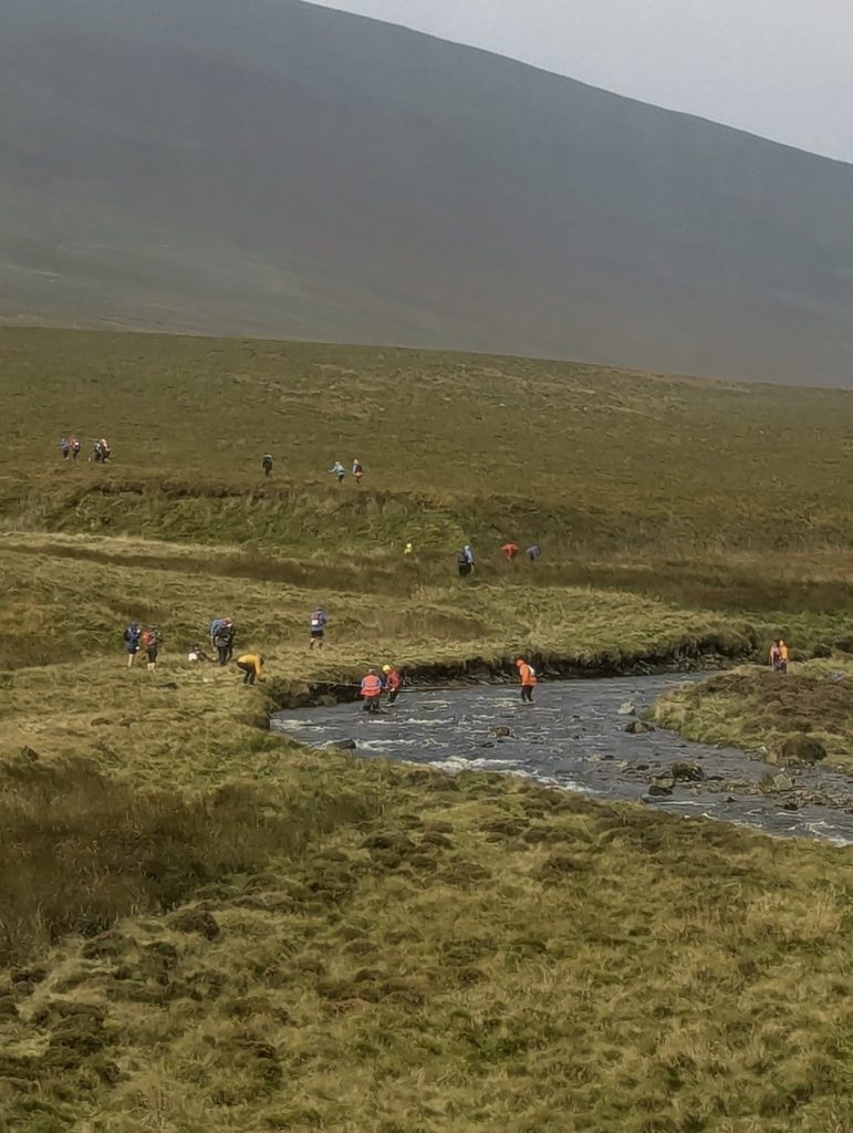 Participants wading through a cold, rushing river crossing supported by marshals on course.