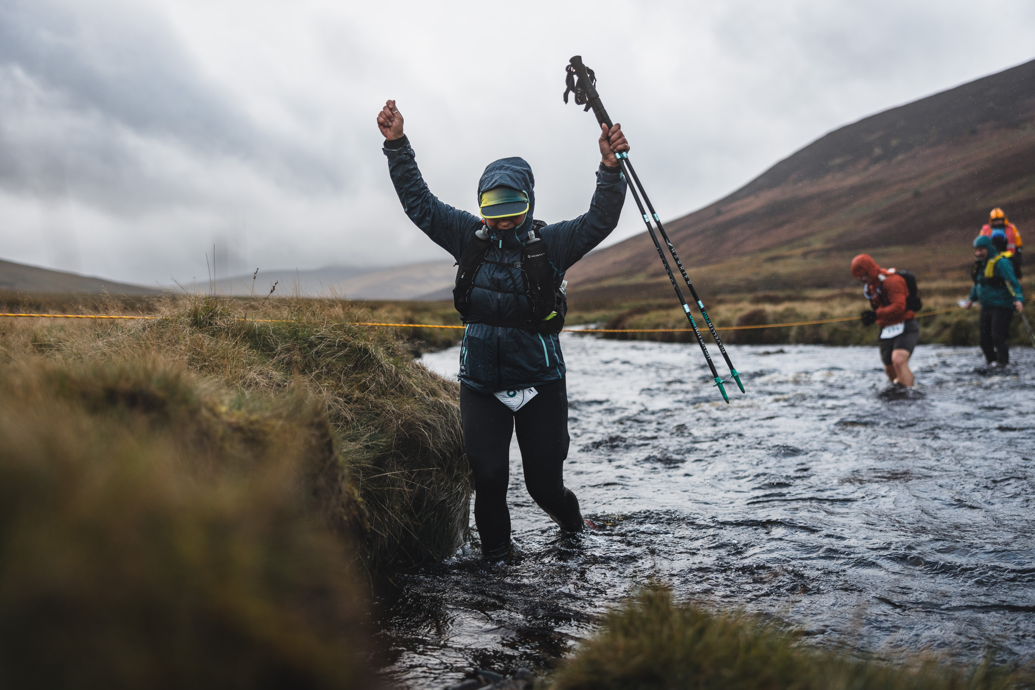 Trail runner raising arms in celebration while wading through a freezing, knee-deep river crossing with trekking poles.