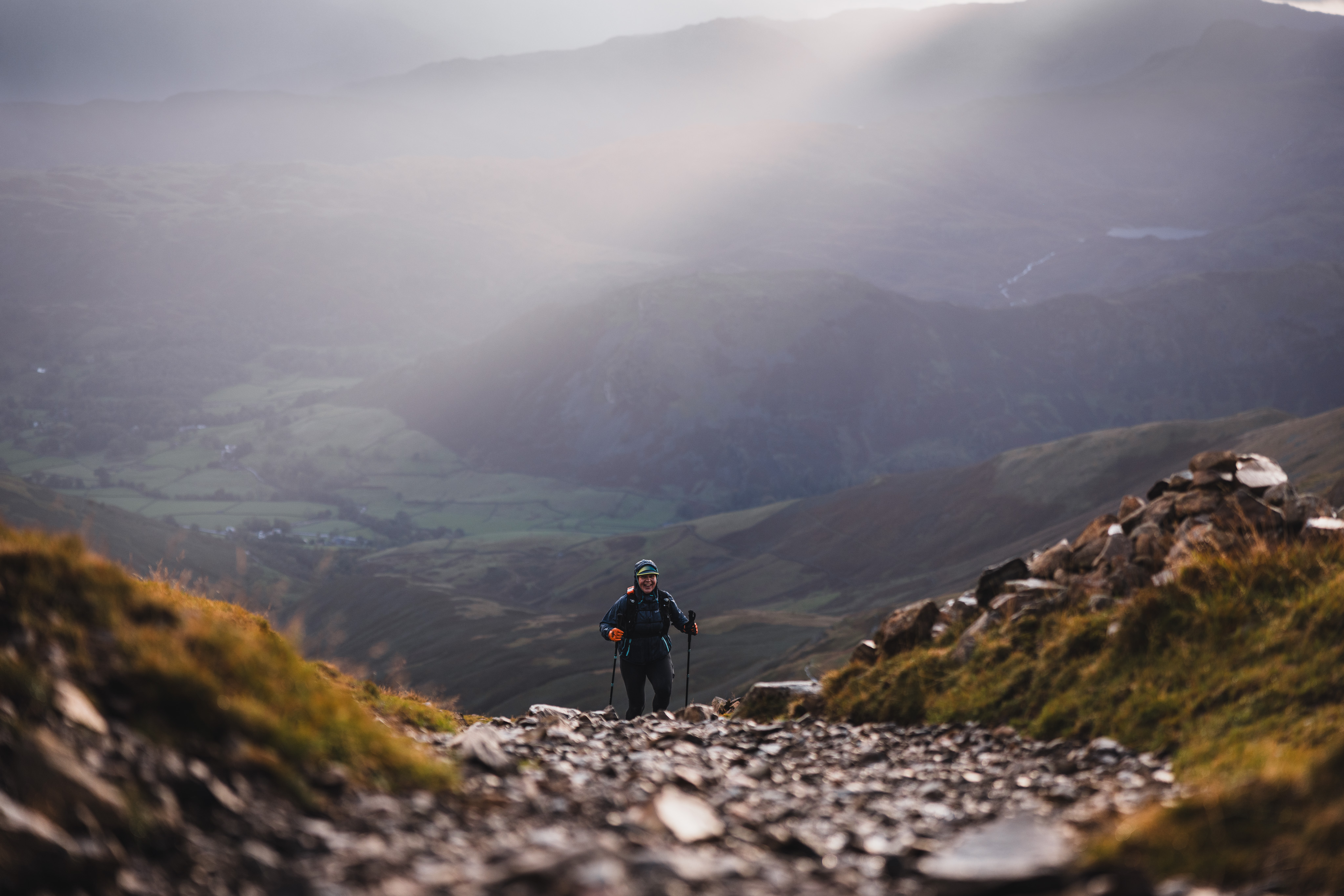 Ultra runner using trekking poles to ascend a steep, rocky mountain path bathed in dramatic valley sunlight.