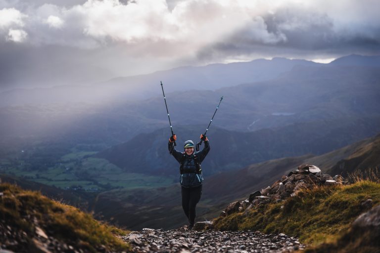 Trail runner raising trekking poles in triumph at a fell summit with dramatic mountain clouds behind.