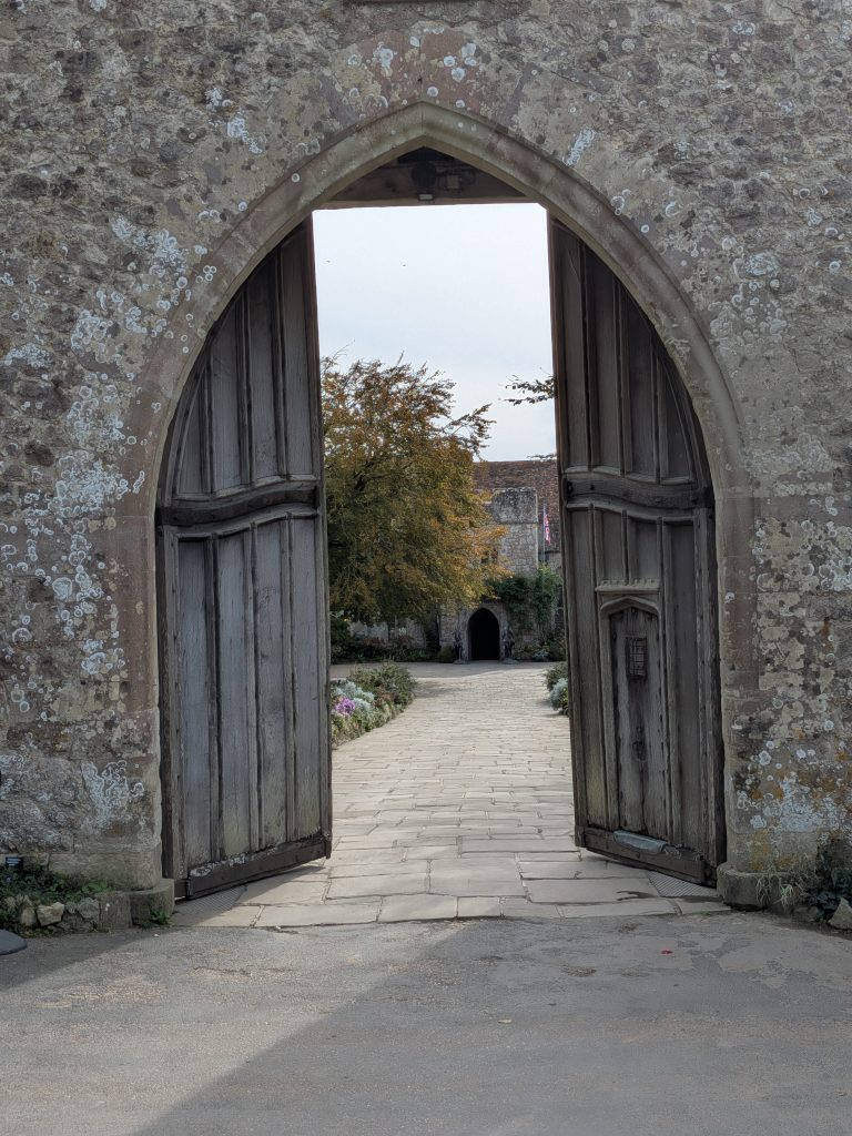 Massive, weathered wooden double doors set within a stone gothic archway, opening to reveal a paved pathway leading into the historic courtyard of Lympne Castle.