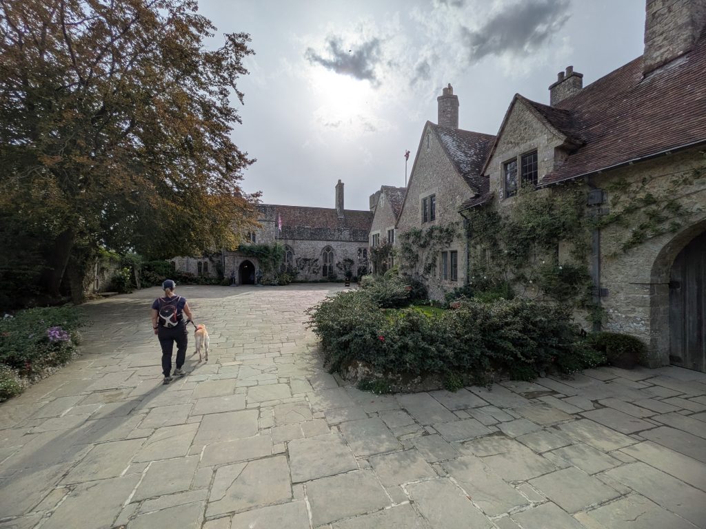 Syla wearing a blue trail pack, walking yellow Labrador cross Poppy on a lead across a wide flagstone courtyard toward the historic stone buildings of Lympne Castle.
