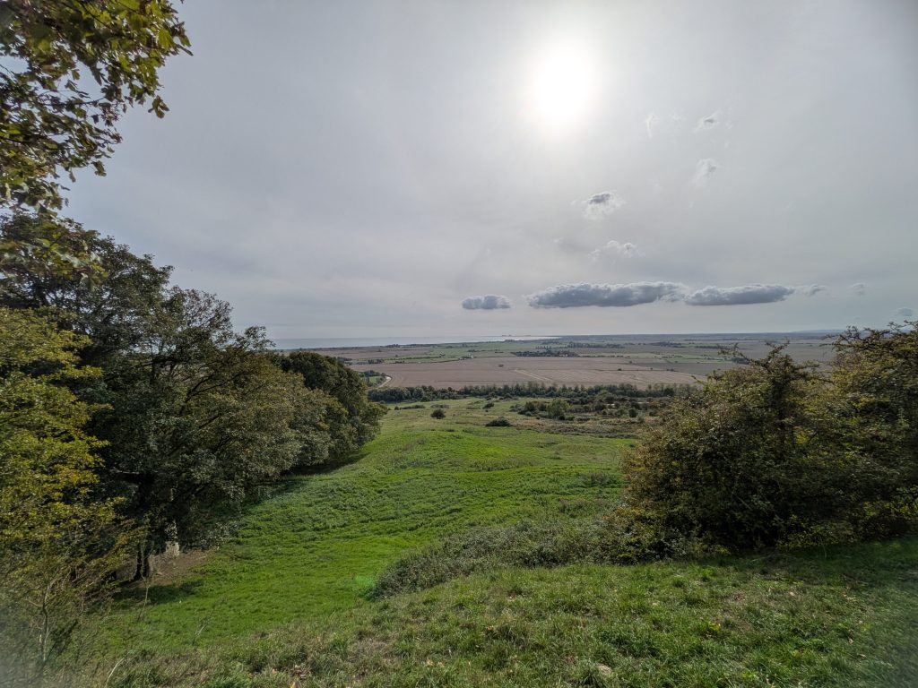 Panoramic view from a high grassy escarpment looking down over flat agricultural fields toward the distant Romney Marsh coastline under a bright, hazy sky.