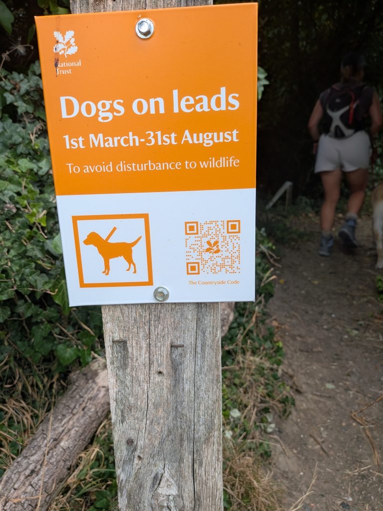 An orange National Trust sign reading "Dogs on leads 1st March-31st August" with a hiker and dog walking past in the background.