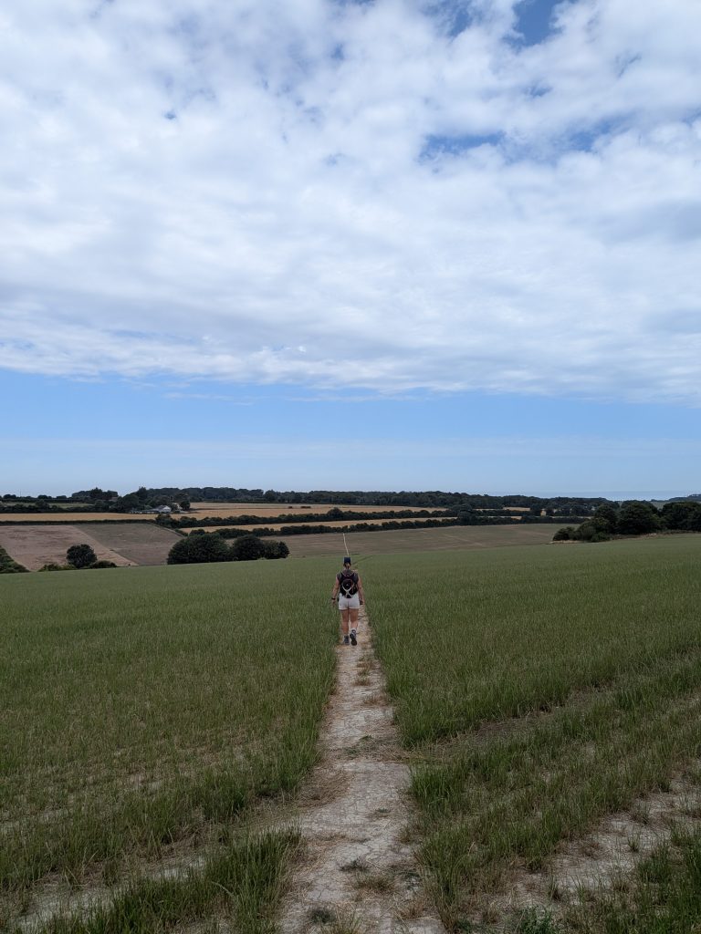 A female hiker with a backpack walking away down a straight, chalky path through a large green agricultural field near the Kent coast.